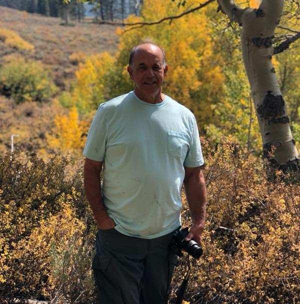 Robert Cherenson wearing a teal shirt standing in some brush holding a camera. 